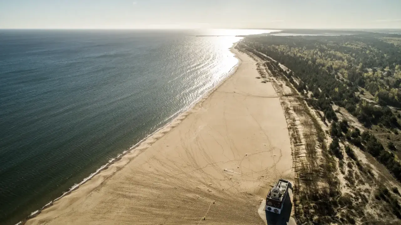 Szeroka, piaszczysta plaza Stogi rozciąga się wzdłuż spokojnego morza. W oddali widać las i budynek na plaży.