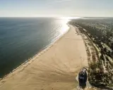 Szeroka, piaszczysta plaza Stogi rozciąga się wzdłuż spokojnego morza. W oddali widać las i budynek na plaży.
