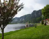 Kwiaty na drzewie i zielona trawa nad rzeką, w tle skaliste góry i miasteczko. Idealne miejsce na spacer, jak z **bastei cennik** wycieczki.