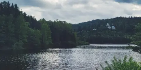 Spokojna tafla wody jeziora, otoczona gęstym lasem. Na wzgórzu majaczy pałac, przypominający scenerię z Wisły Kubalonki.