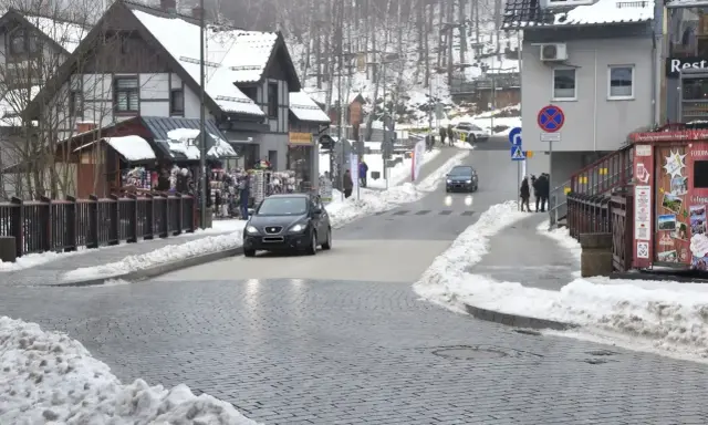 Zimowy deptak w Szklarskiej Porębie. Samochody jadą po ośnieżonej drodze, a po bokach widać budynki i stragany.