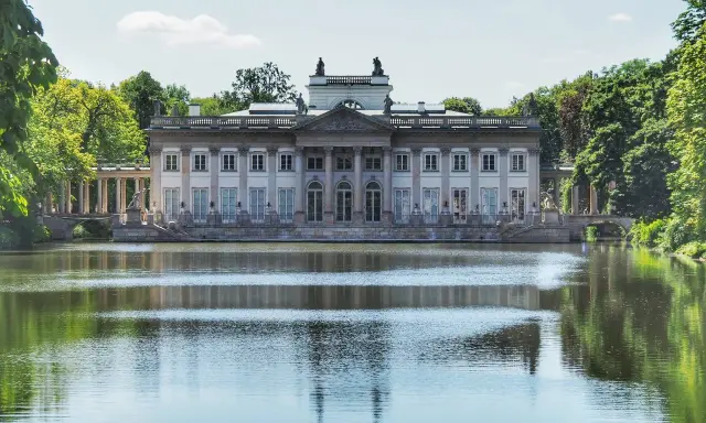 Pałac na Wodzie w Łazienkach Królewskich odbija się w spokojnej tafli jeziora, otoczony zielenią drzew.