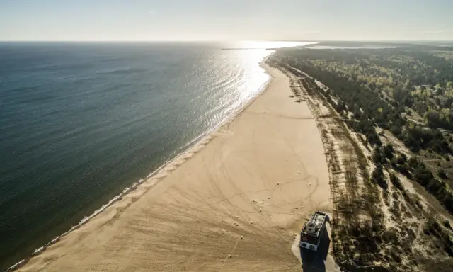 Szeroka, piaszczysta plaza Stogi rozciąga się wzdłuż spokojnego morza. W oddali widać las i budynek na plaży.