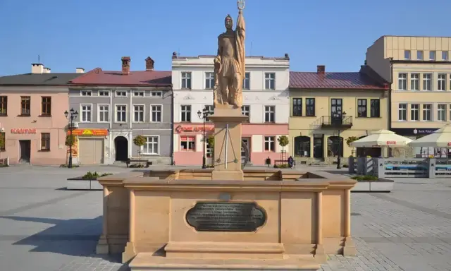 Pomnik na rynku w Żywcu. Kamienna fontanna z rzeźbą żołnierza na tle kolorowych kamienic.