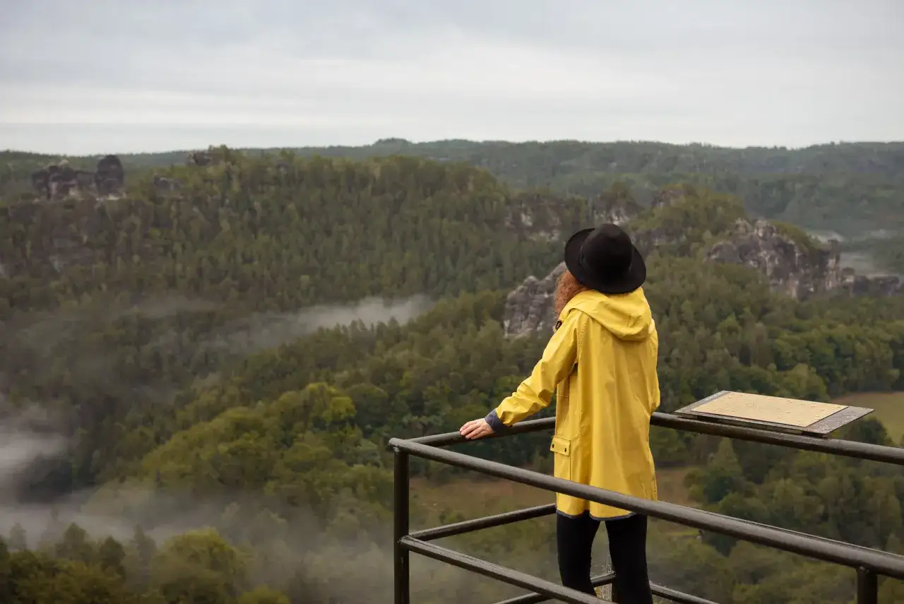 Kobieta w żółtej kurtce i czarnym kapeluszu podziwia widok na skaliste bastei. Obok niej znajduje się tablica informacyjna, przypominająca cennik.